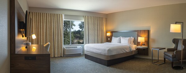 Ein Hotelzimmer mit Bett, Nachttischen, einem Schreibtisch, einem Sessel und Blick auf Bäume durch das Fenster.