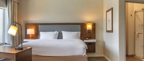 A hotel room with a large bed, two bedside lamps, a wooden headboard, a small bedside table, a waste bin, and a door leading to another room.