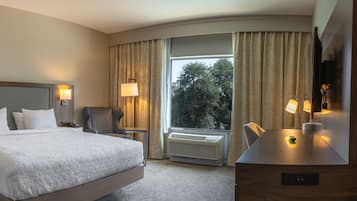 A hotel room with a large bed, two bedside tables with lamps, a chair, a desk with a lamp, a large window with curtains, and an air conditioning unit.