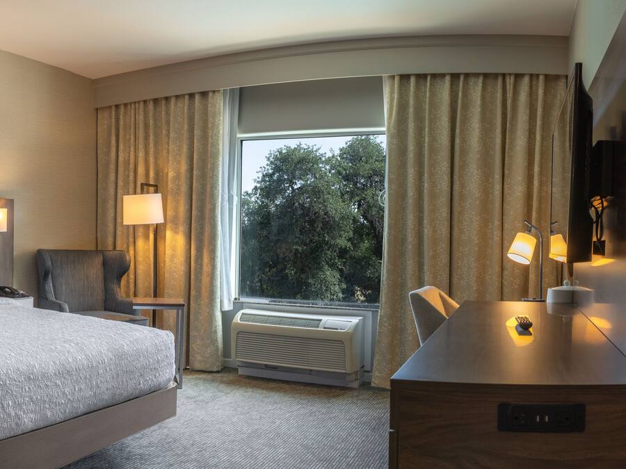 A hotel room with a large bed, two bedside tables with lamps, a chair, a desk with a lamp, a large window with curtains, and an air conditioning unit.