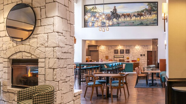 A cozy restaurant with a stone fireplace, wooden tables, and houndstooth chairs. - Hampton Inn & Suites Boerne (Boerne)
