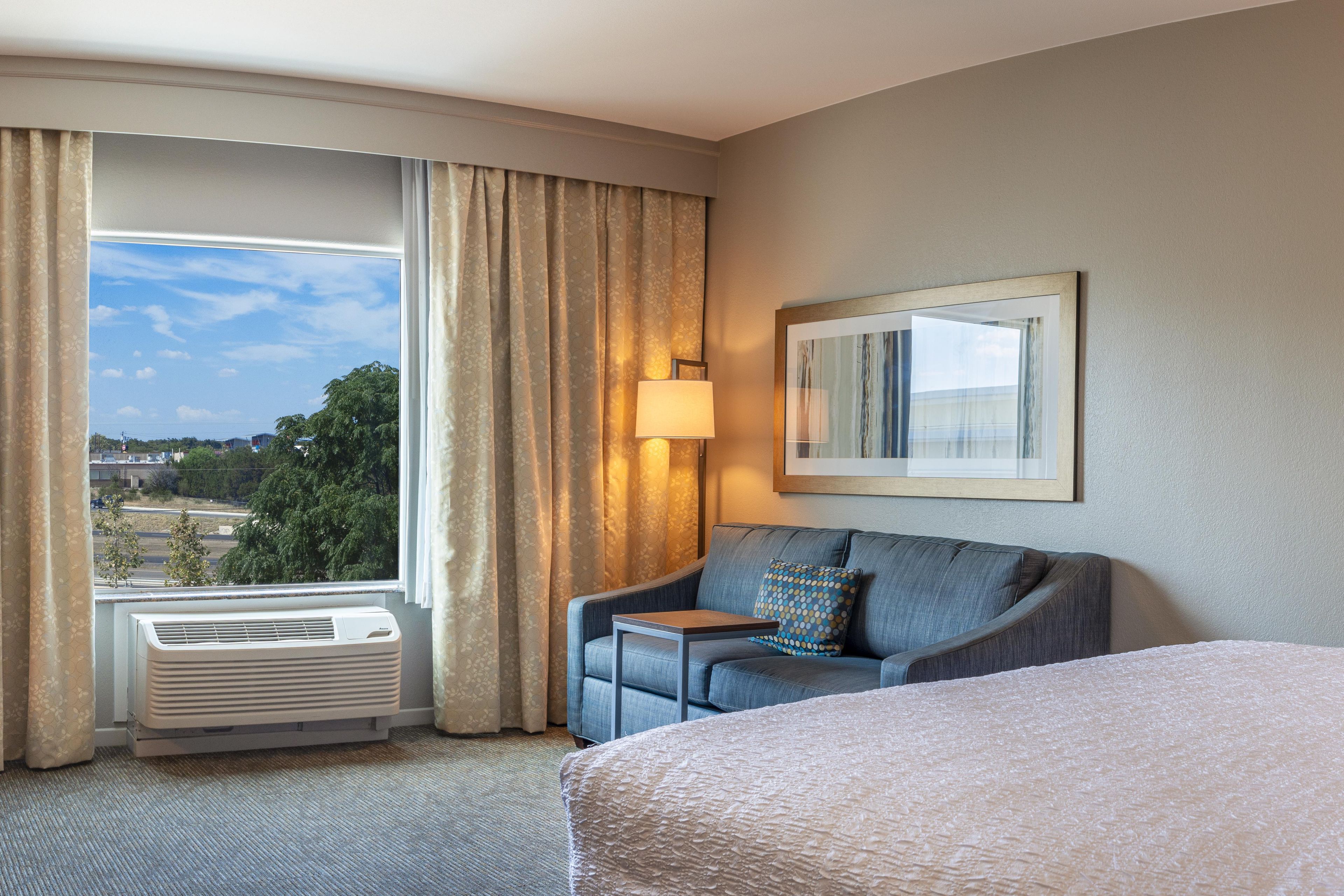 A hotel room with a bed, a sofa, a table, a lamp, and a window with curtains.
