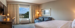 A hotel room with a bed, a desk, a chair, a sofa, a window with curtains, a lamp, and a painting on the wall. - Hampton Inn & Suites Boerne (Boerne)
