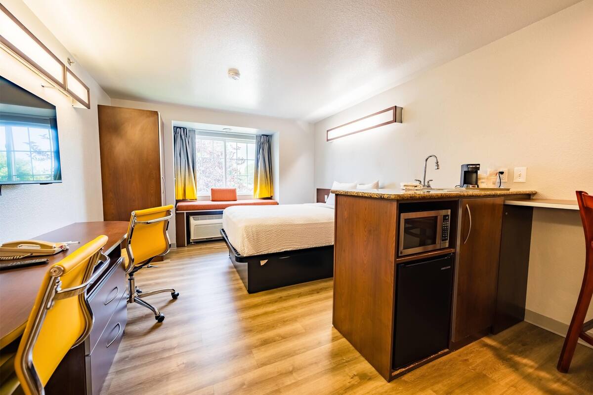 A compact hotel room with a wooden desk, a single bed, a kitchenette, and a window with curtains.