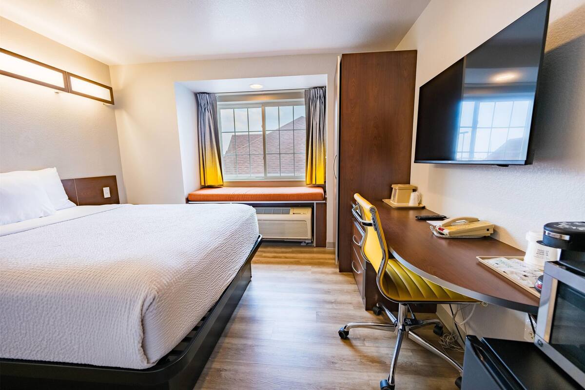 A hotel room with a bed, a desk with a chair, a flat-screen TV, and a window with curtains.