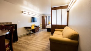 A hotel room with a wooden floor, a sofa, a desk with a chair, a television mounted on the wall, and a window with curtains.