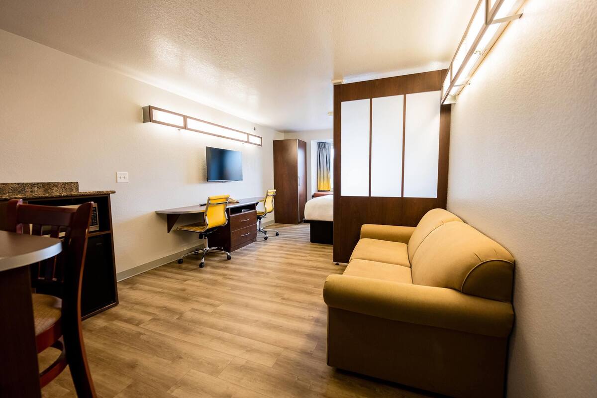 A hotel room with a wooden floor, a sofa, a desk with a chair, a television mounted on the wall, and a window with curtains.