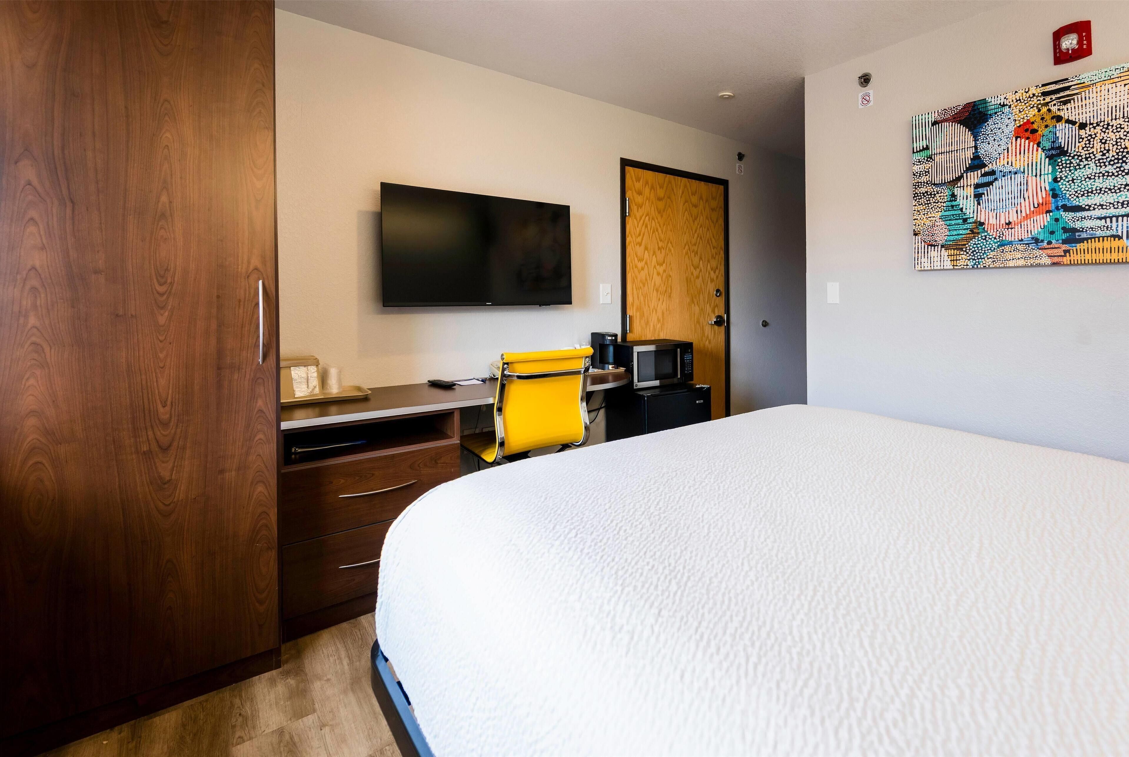 A hotel room with a large bed, a desk with a chair, a flat-screen TV, a coffee maker, and a wooden wardrobe.