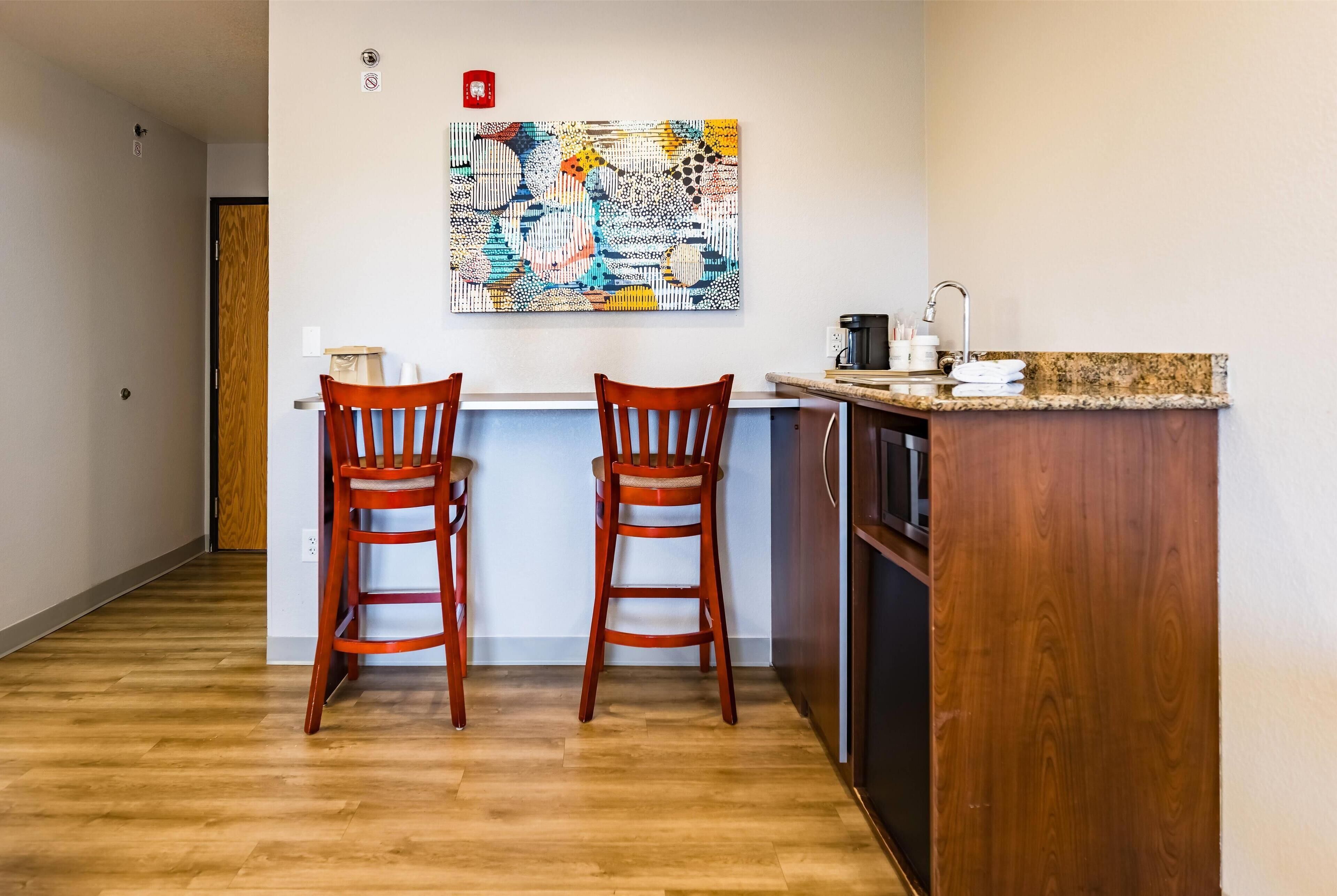 A small kitchen with two wooden chairs, a granite countertop, and a colorful abstract painting on the wall.