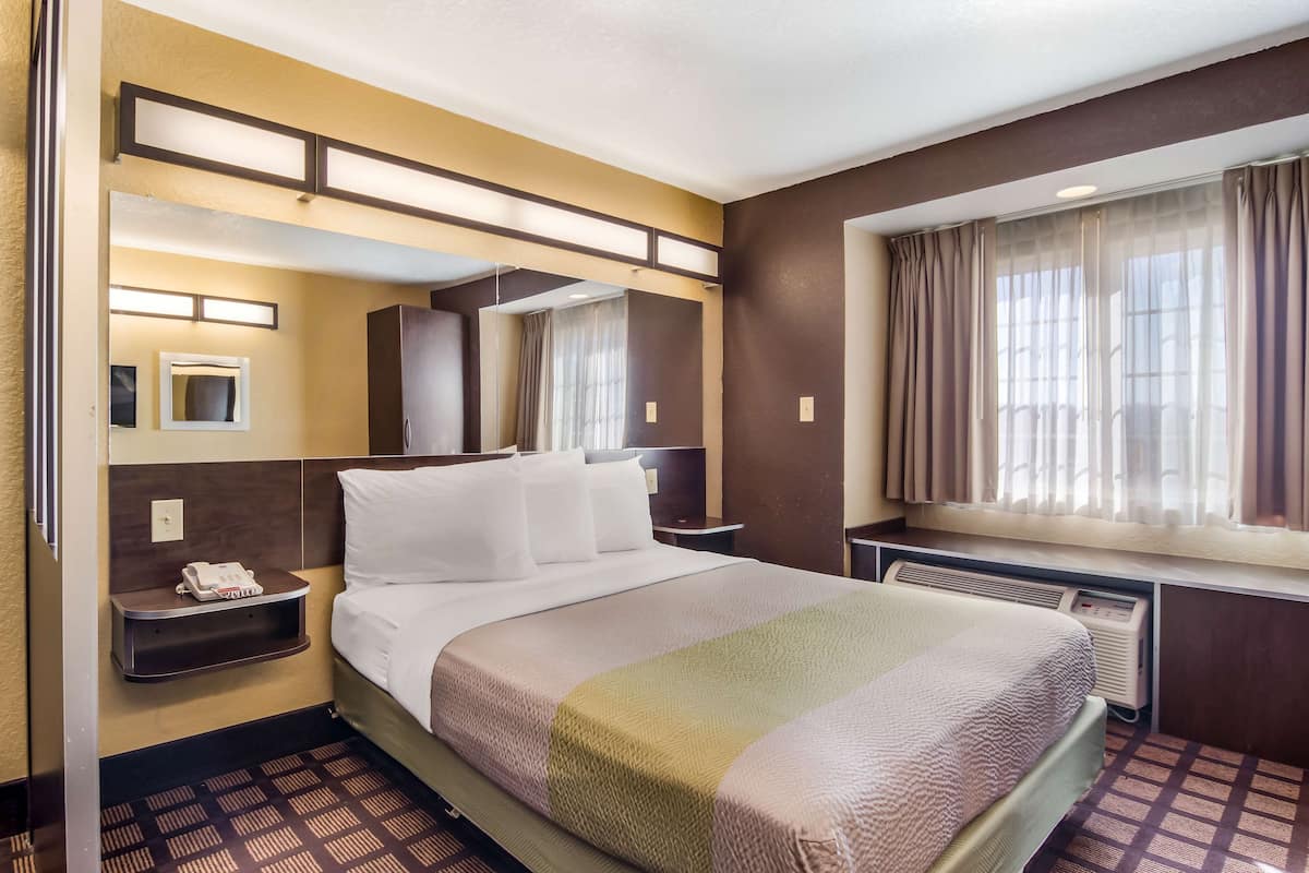 A hotel room with a bed, bedside tables, a mirror, a window with curtains, and a wall-mounted heater.