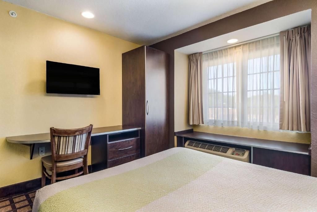 Habitación de hotel con cama, escritorio, silla, televisor y ventana con cortinas.