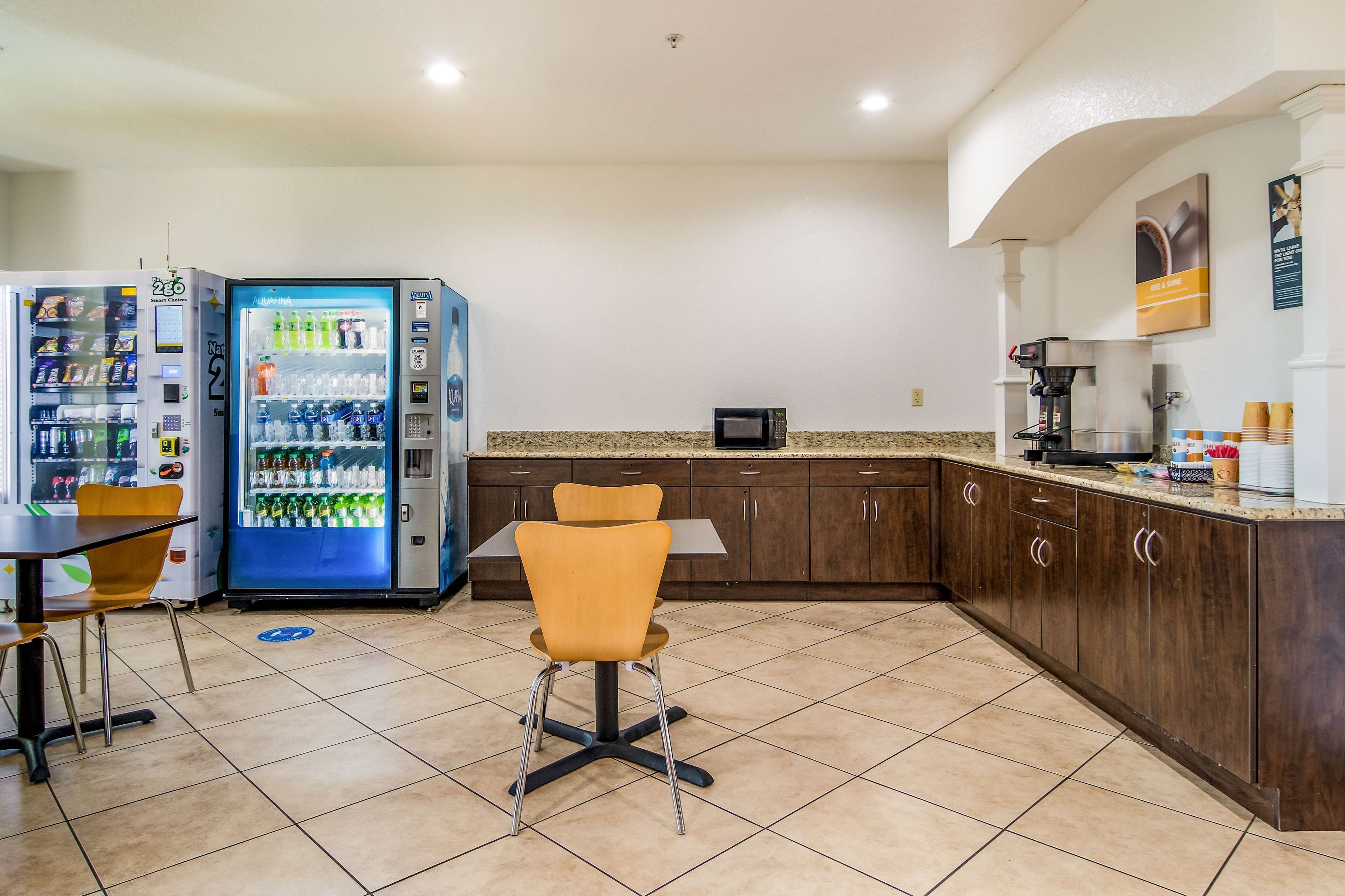 A clean, modern kitchen with a microwave, coffee machine, and a vending machine. There is a table and a chair in the center.