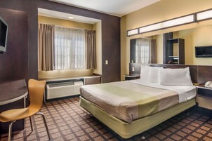 Habitación de hotel con cama, una silla, un escritorio, televisor, espejo y ventana con cortinas.