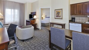 Habitación de hotel con cama, escritorio, silla, sofá, televisor, lámpara, un cuadro y una cocina equipada con cafetera y lavavajillas.