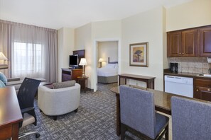 A hotel room with a bed, a desk, a chair, a sofa, a television, a lamp, a painting, and a kitchen area with a coffee maker and dishwasher. - Staybridge Suites Lansing - Okemos by IHG (Okemos)