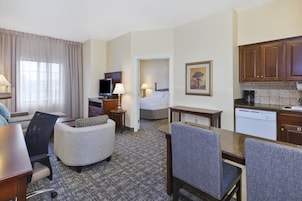 Habitación de hotel con cama, escritorio, silla, sofá, televisor, lámpara, un cuadro y una cocina equipada con cafetera y lavavajillas.