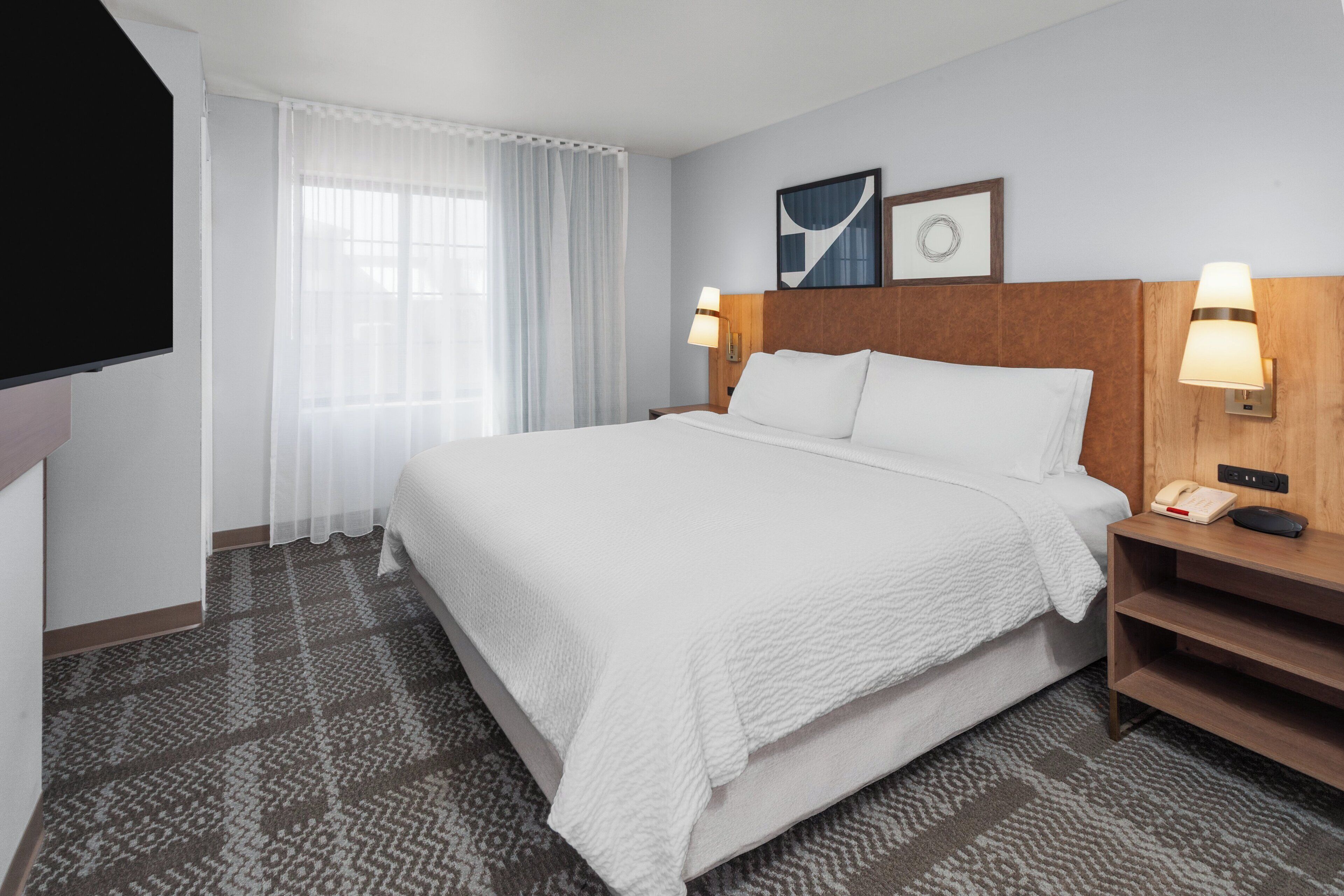 Une chambre d’hôtel avec un grand lit, un téléviseur à écran plat, une table de chevet avec une lampe et une fenêtre avec des rideaux légers.