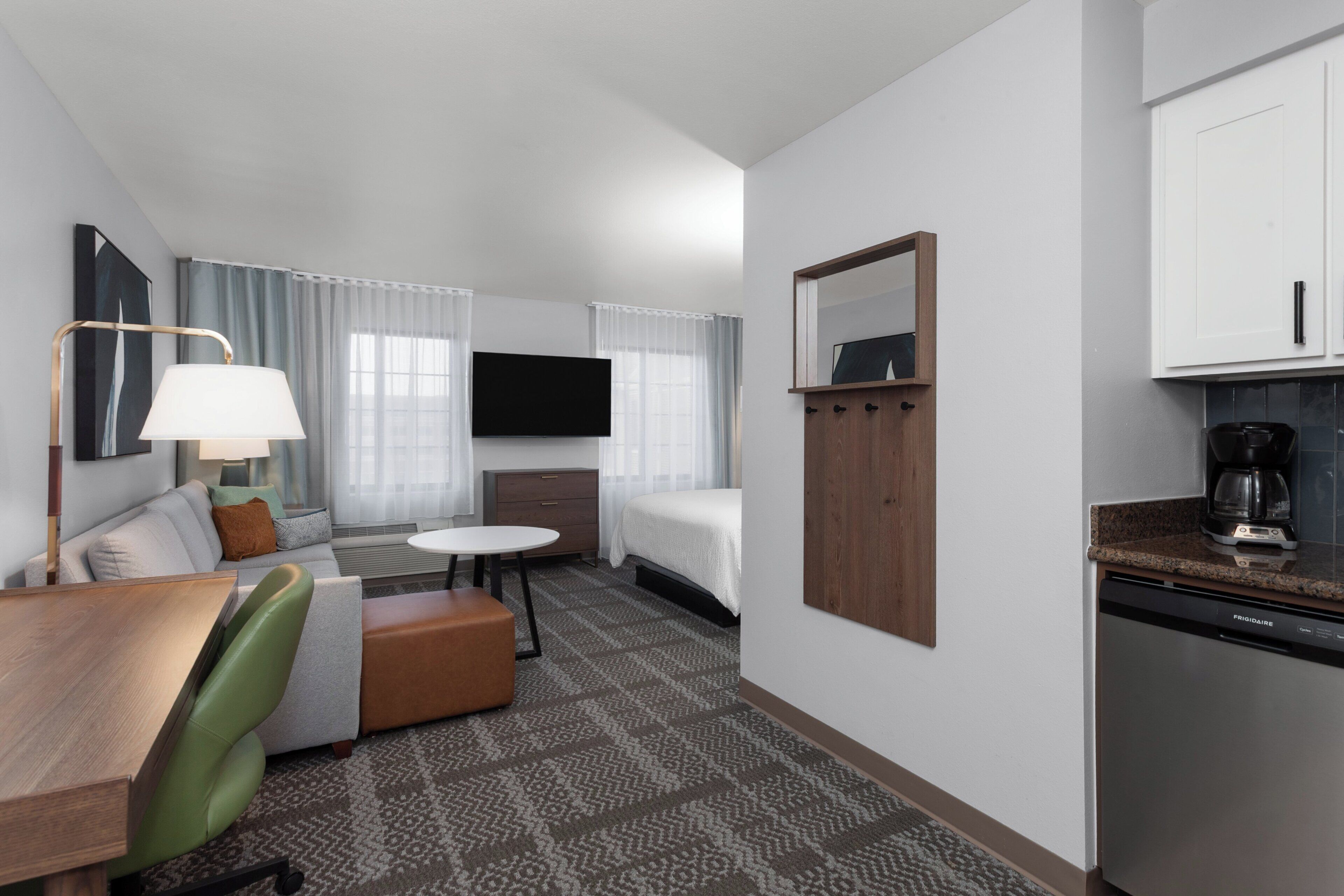 A hotel room with a bed, a desk, a chair, a TV, a coffee maker, and a kitchen area.