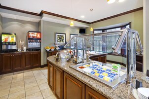 Una barra de desayuno con servicio tipo buffet, que incluye una máquina de café, dispensador de zumo y diversas opciones de alimentos.