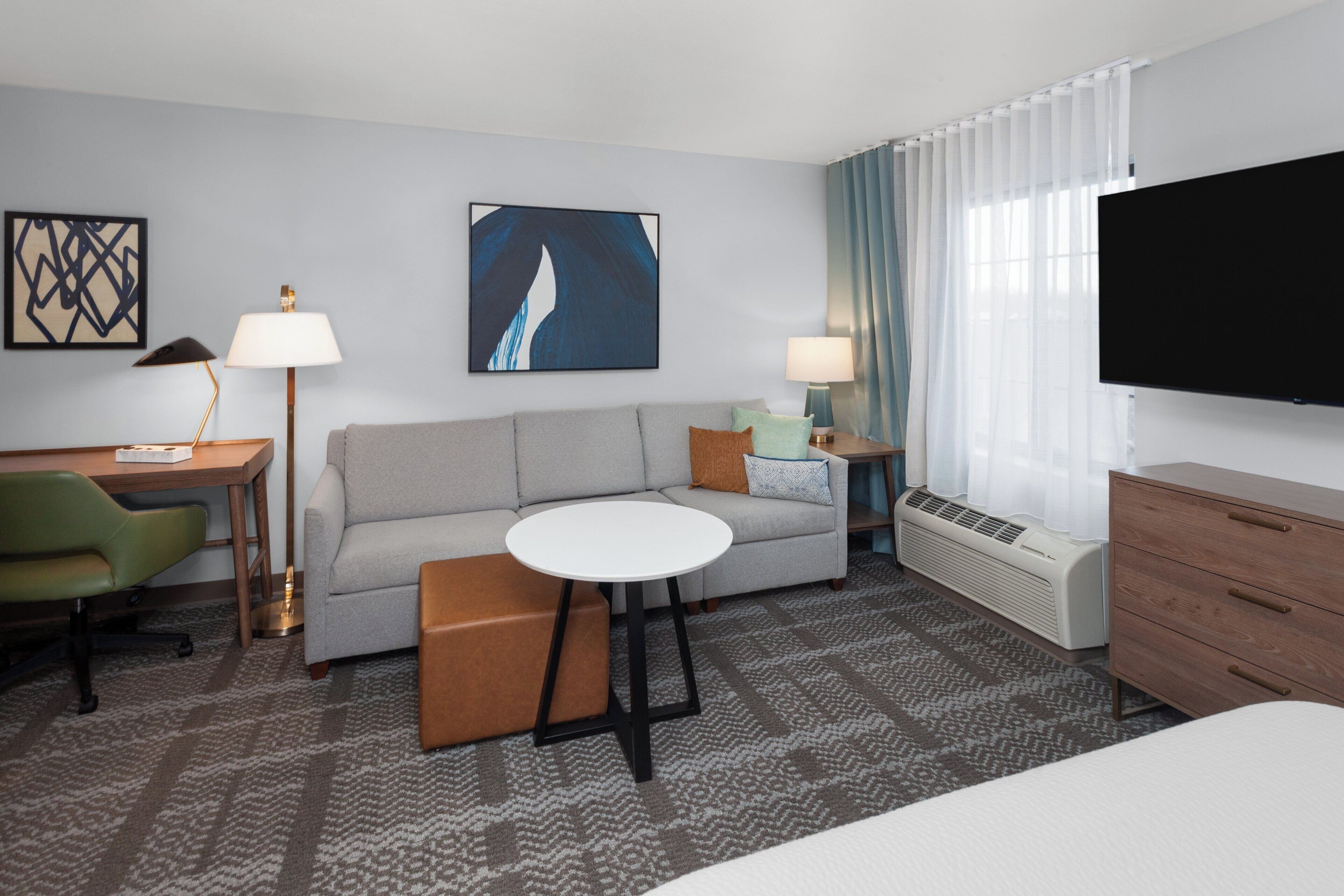 A hotel room with a grey sofa, a small round table, a green chair, a TV, and a heater.