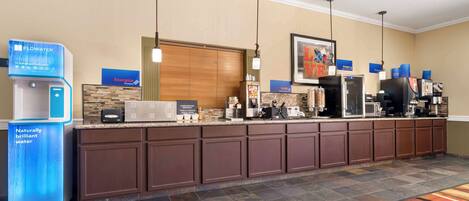 Zona de recepción de hotel con dispensador de agua, máquina de café y mostrador con diversos servicios.