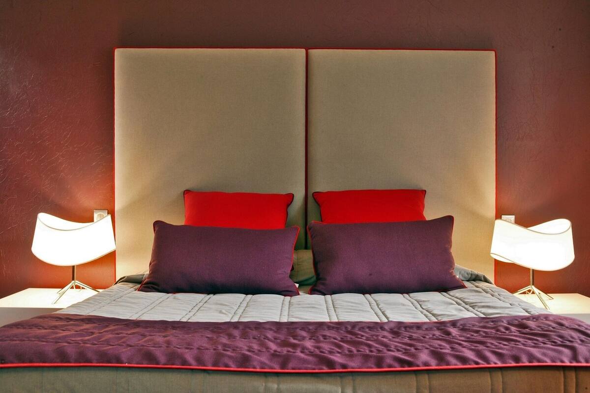 A bed with a headboard, two red pillows, two purple pillows, and a purple blanket.