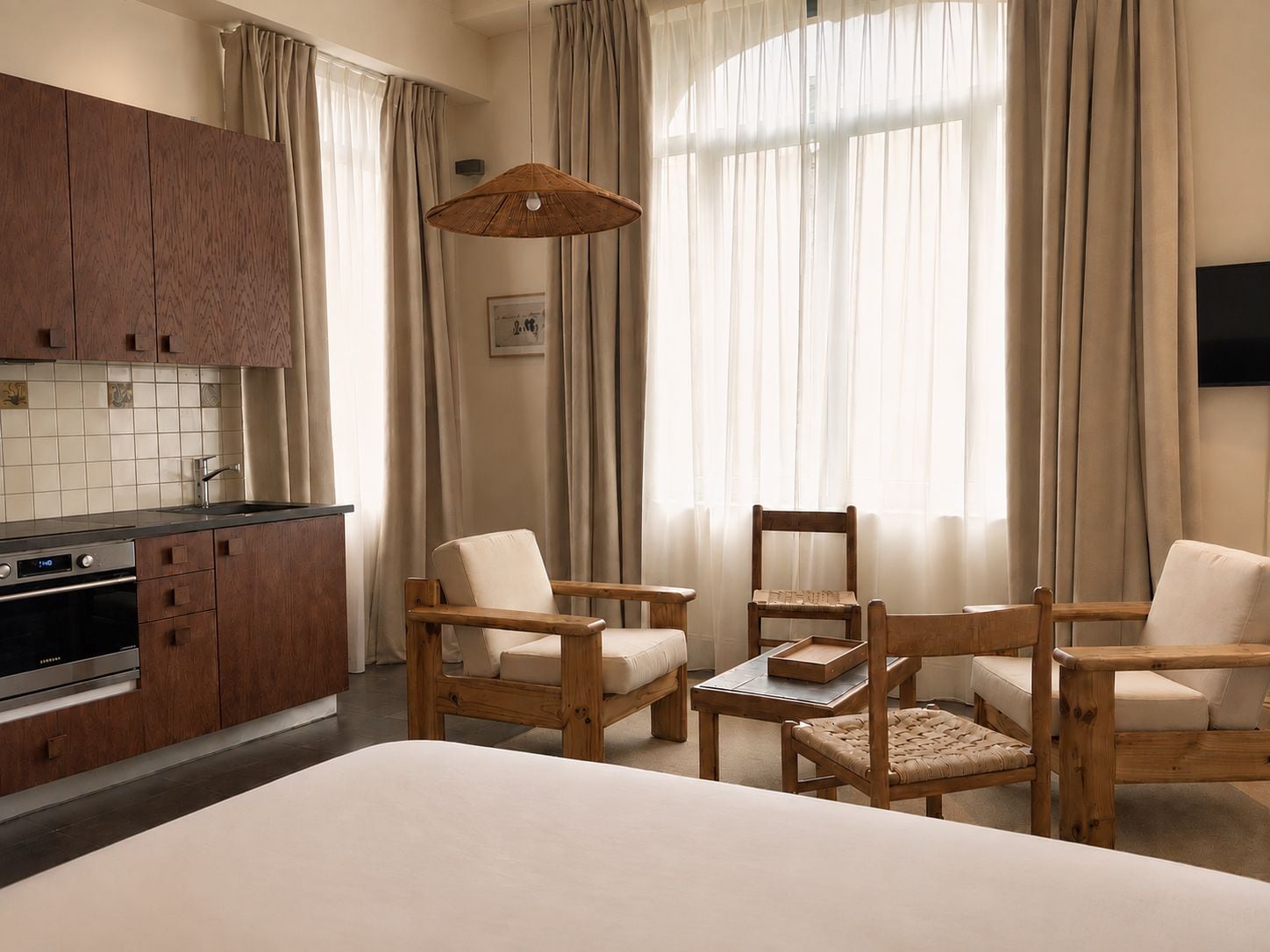 Ein Hotelzimmer mit Küchenzeile, zwei Holzstühlen, einem Bett und großen Fenstern mit Vorhängen.