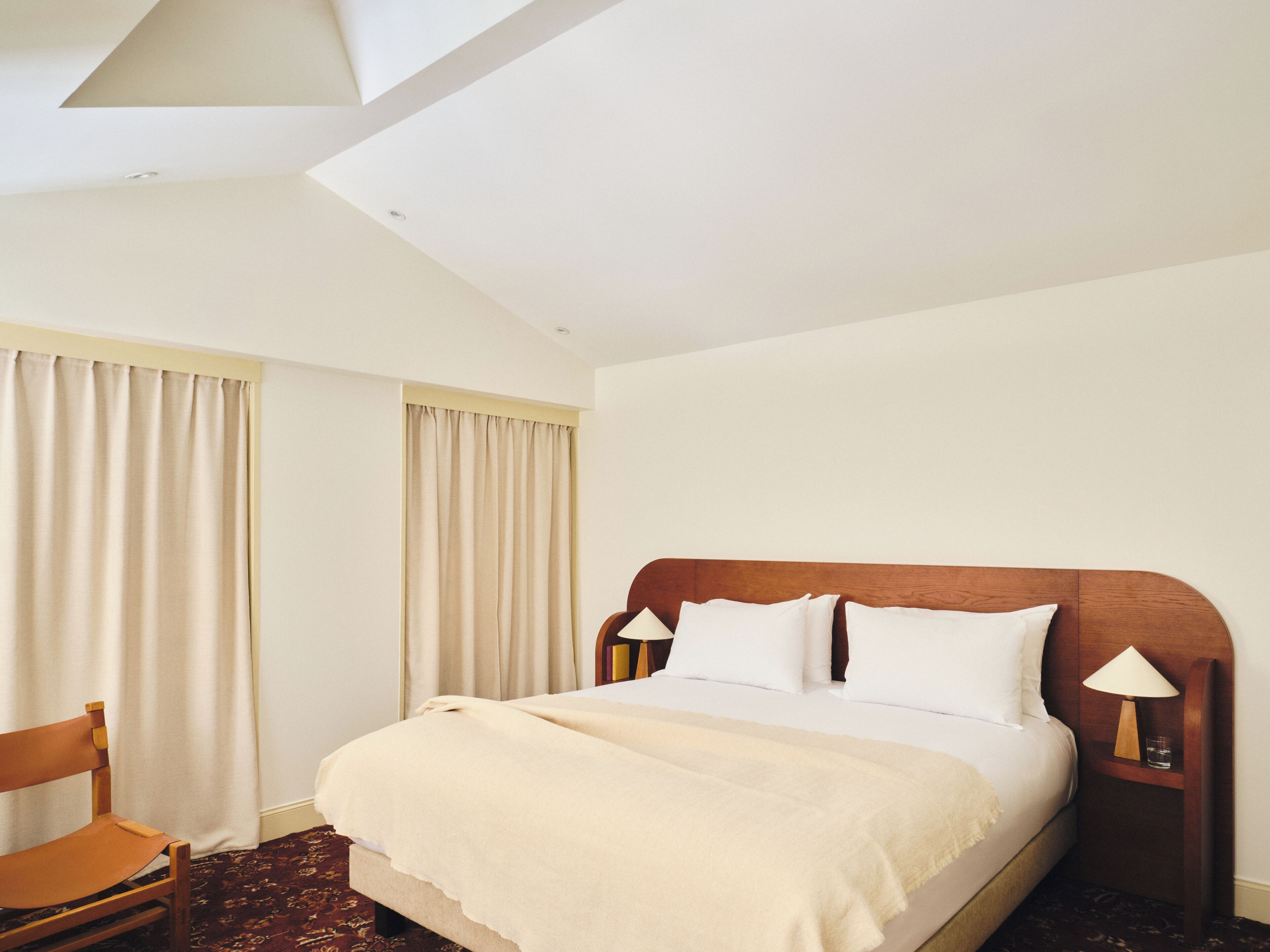 A hotel room with a bed, a chair, a nightstand with a lamp, and a window with curtains.
