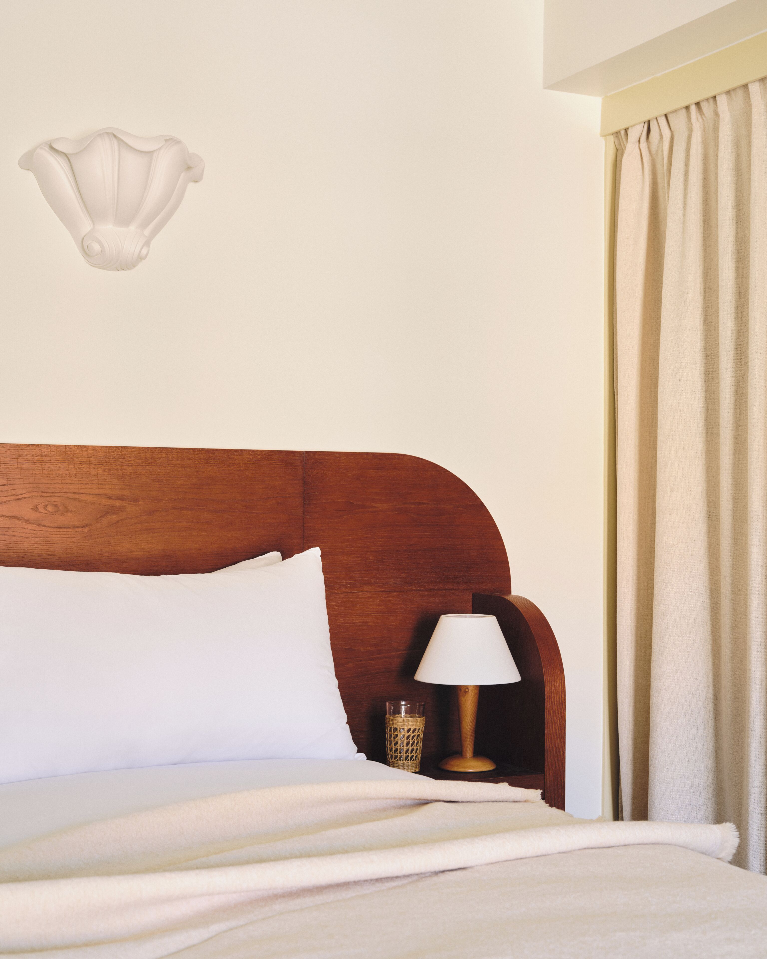 Een bed met witte beddengoed, een houten hoofdbord, een bedlampje en een wandlamp.