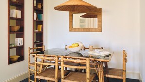 Un coin repas avec une table en verre, des chaises en osier, un luminaire en fibres tressées et des étagères en bois ornées de livres et d’objets décoratifs.