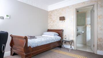 Une chambre à coucher avec un lit en bois, une petite table de chevet, une chaise et une salle de bain visible à travers une porte ouverte.