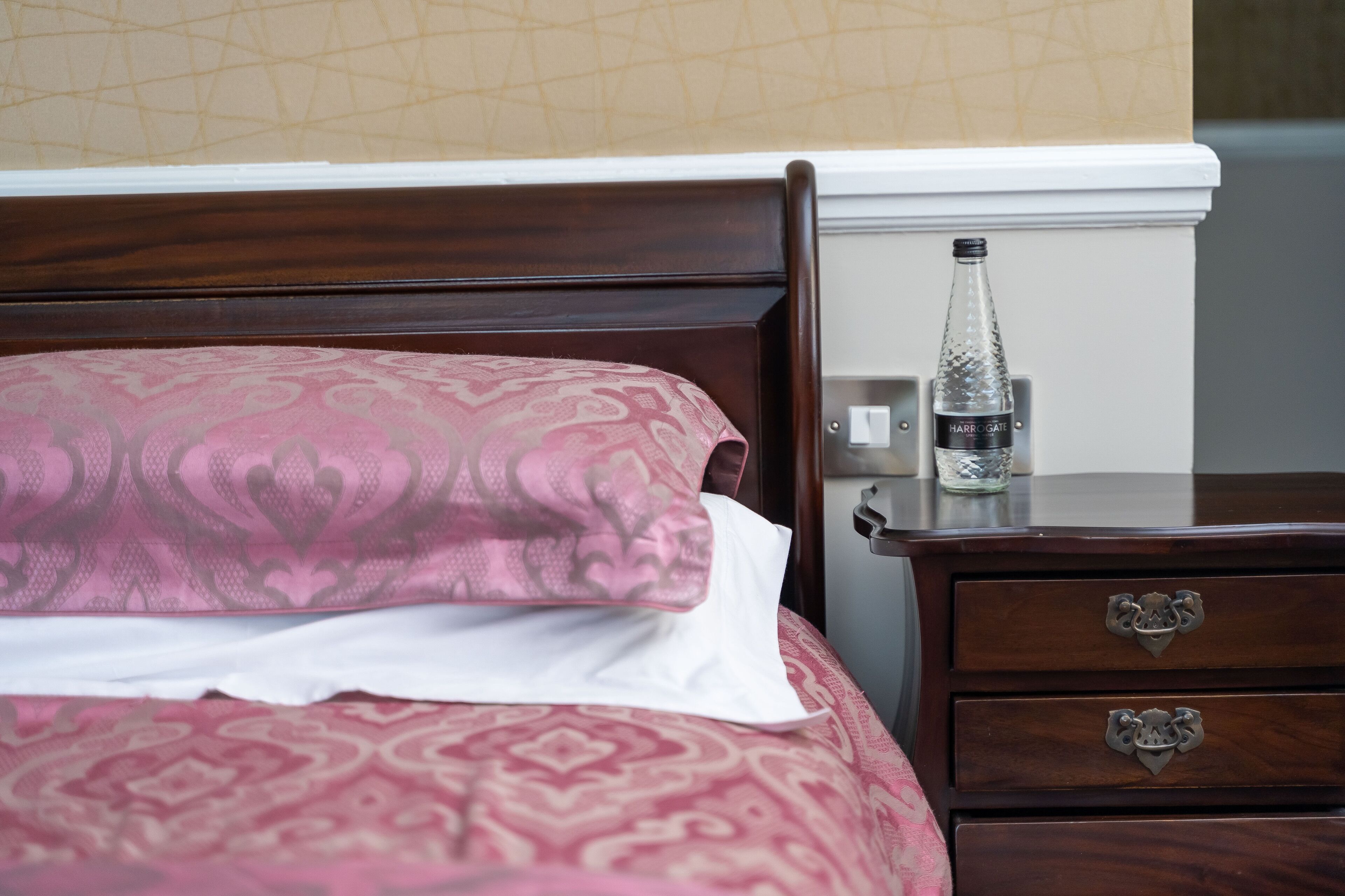 Een net opgemaakt bed met een bedspread met patroon en kussens, een houten nachtkastje met een fles water, en een lade met sierlijke handgrepen.