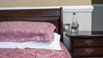 Un lit bien fait, avec une couvre-lit à motifs et des oreillers, une table de chevet en bois sur laquelle se trouve une bouteille d’eau, et un tiroir doté de poignées décoratives.