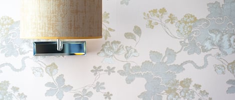 Una lámpara de pared con pantalla texturizada y base metálica, sobre un fondo de papel tapiz con motivos florales.