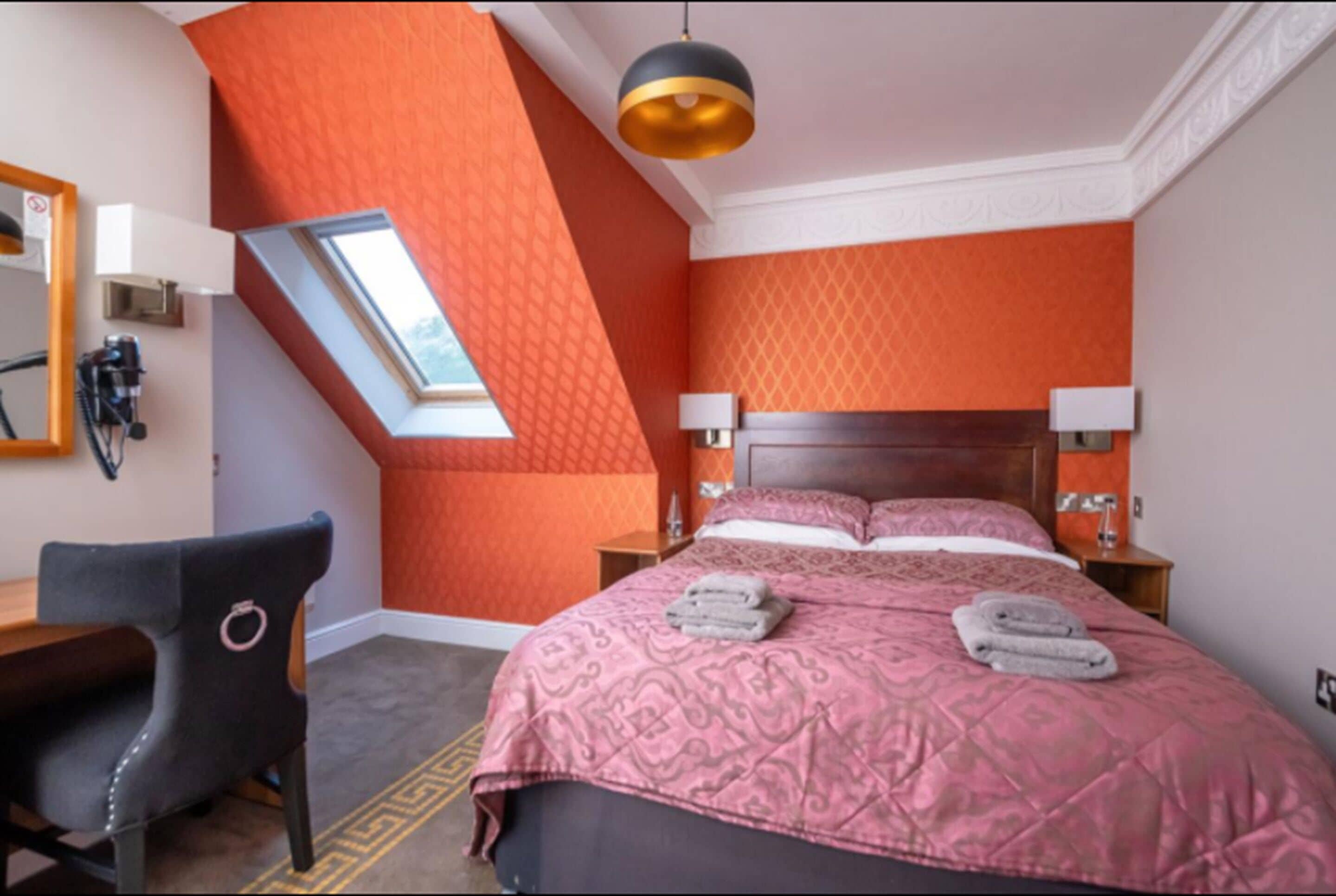 A bedroom with a sloped ceiling, a bed with a patterned bedspread, two bedside lamps, and a desk with a chair.