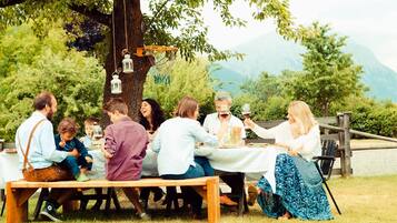 Um grupo de pessoas sentadas em uma mesa de madeira ao ar livre, rodeadas por árvores e montanhas ao fundo.