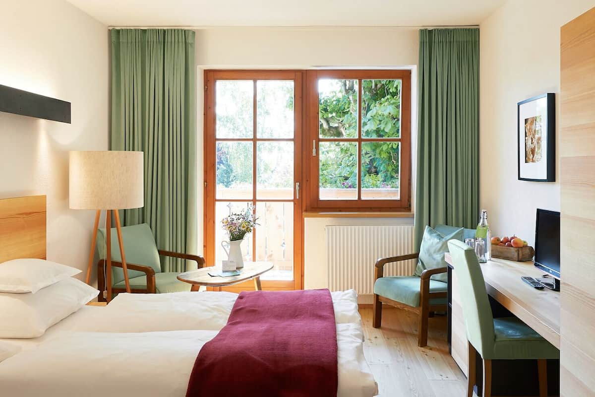 A hotel room with two beds, a desk, a chair, a lamp, a window with curtains, and a radiator.