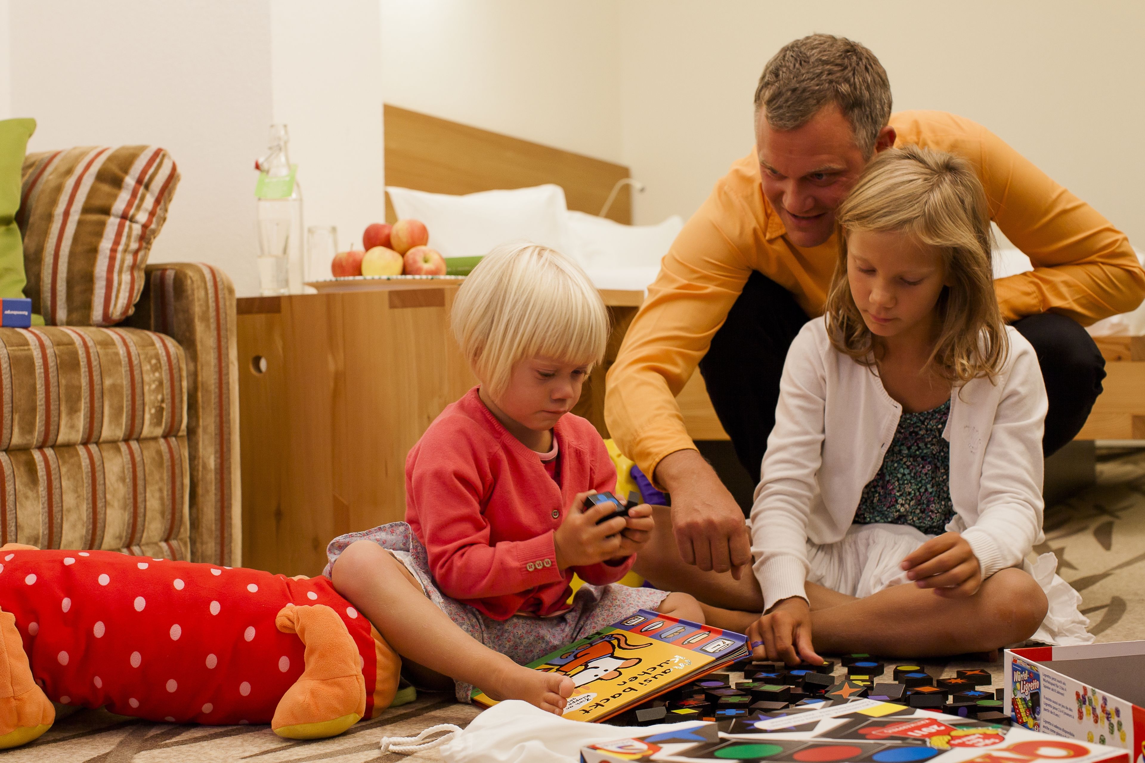 A family of three sitting on the floor in a room with a bed, a wooden cabinet, and a striped armchair.
