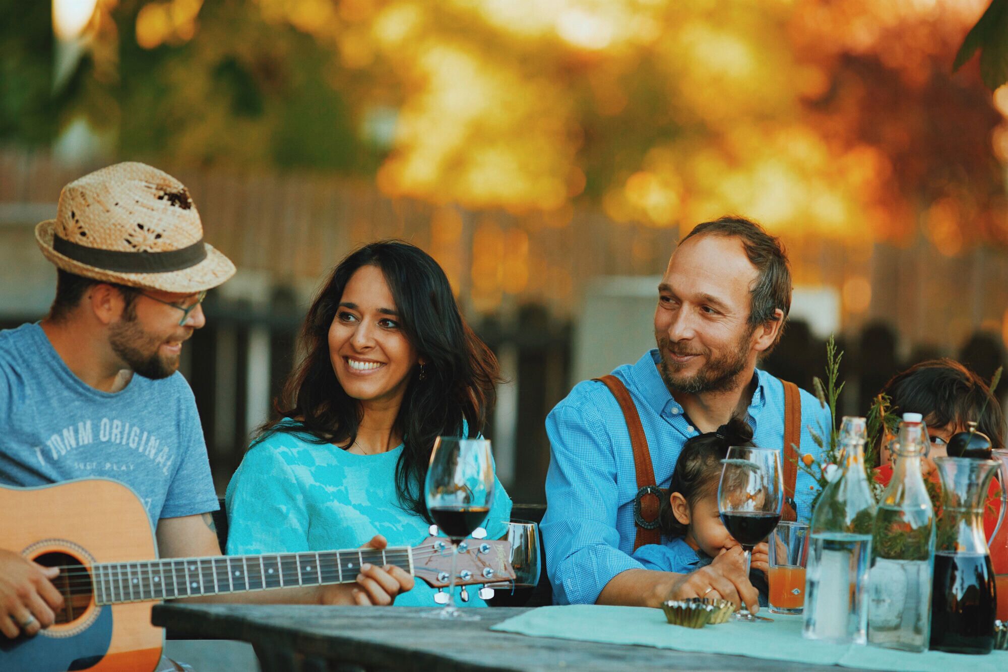 Un gruppo di persone sedute a un tavolo all'aperto, con una chitarra, bicchieri di vino e diverse bottiglie.