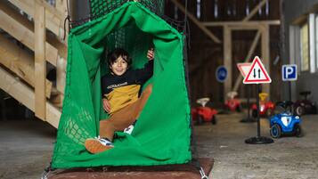 Criança dentro de uma estrutura de rede verde, possivelmente uma área de recreação, com uma rampa de madeira e veículos de brinquedo ao fundo.