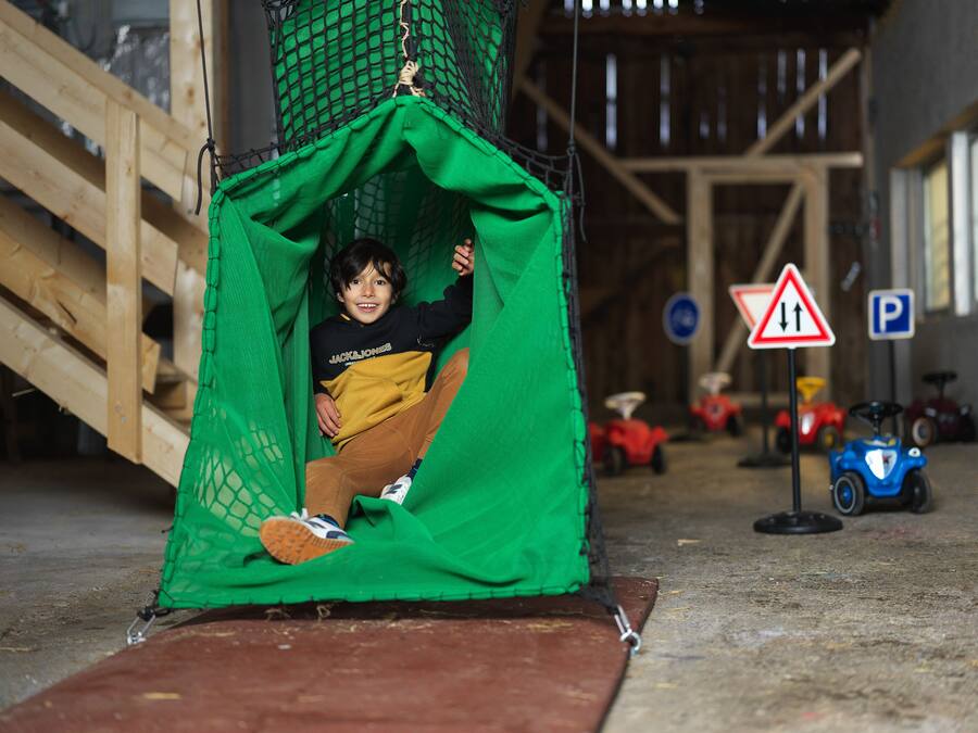 Criança dentro de uma estrutura de rede verde, possivelmente uma área de recreação, com uma rampa de madeira e veículos de brinquedo ao fundo.