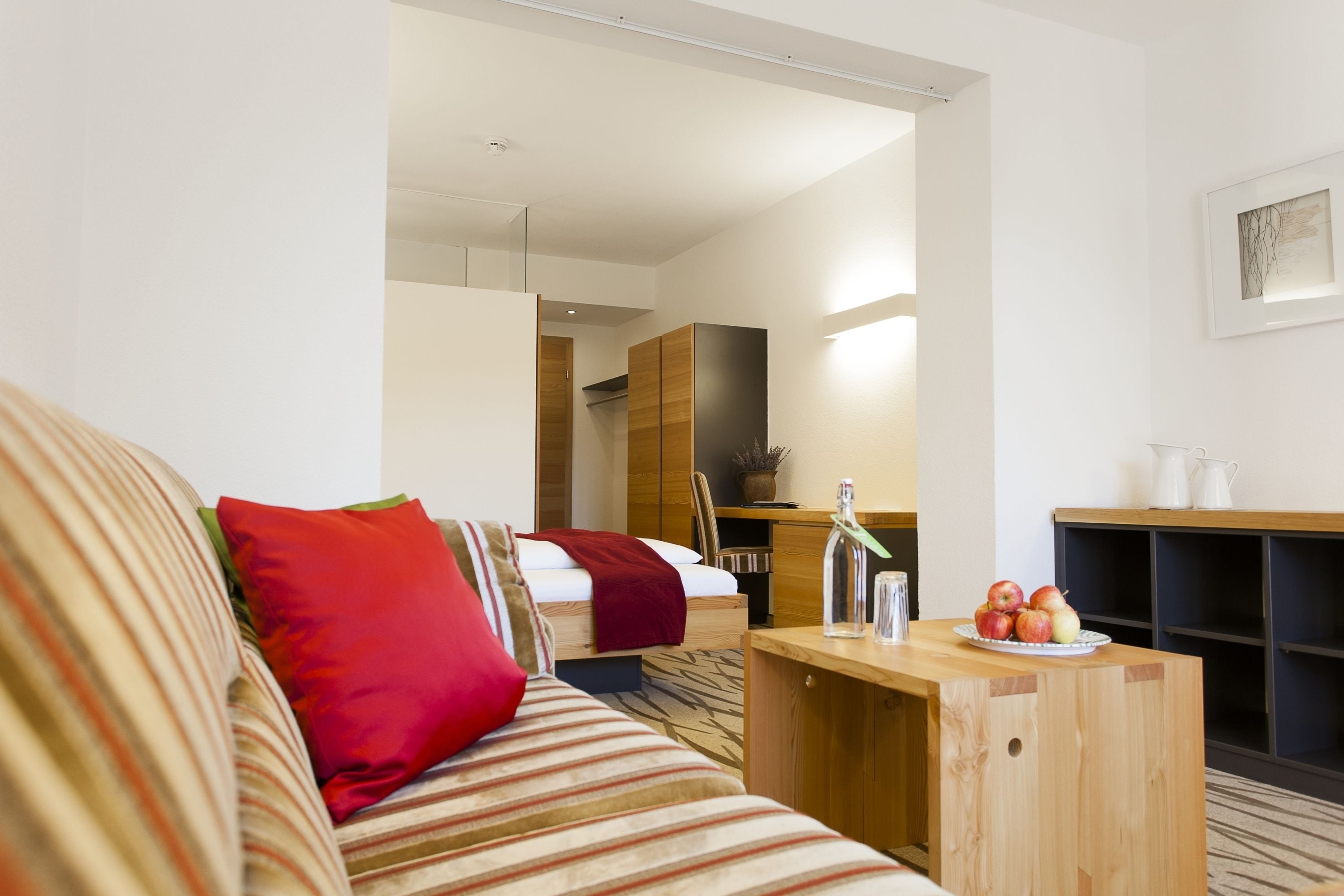 A modern hotel room with a striped sofa, a wooden desk, and a bed with a red pillow.