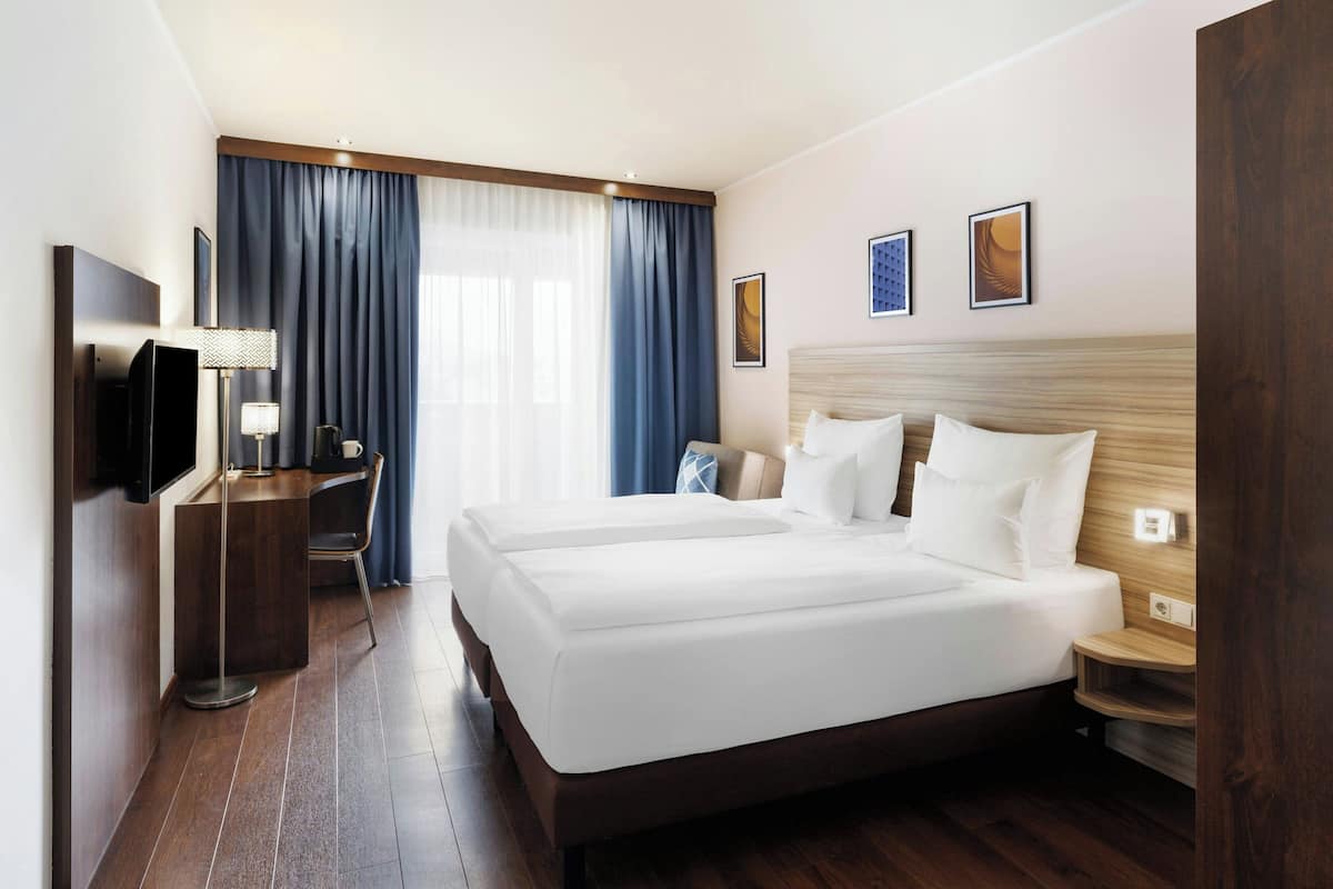 A hotel room with a large bed, a desk with a lamp, a TV, and a window with curtains.