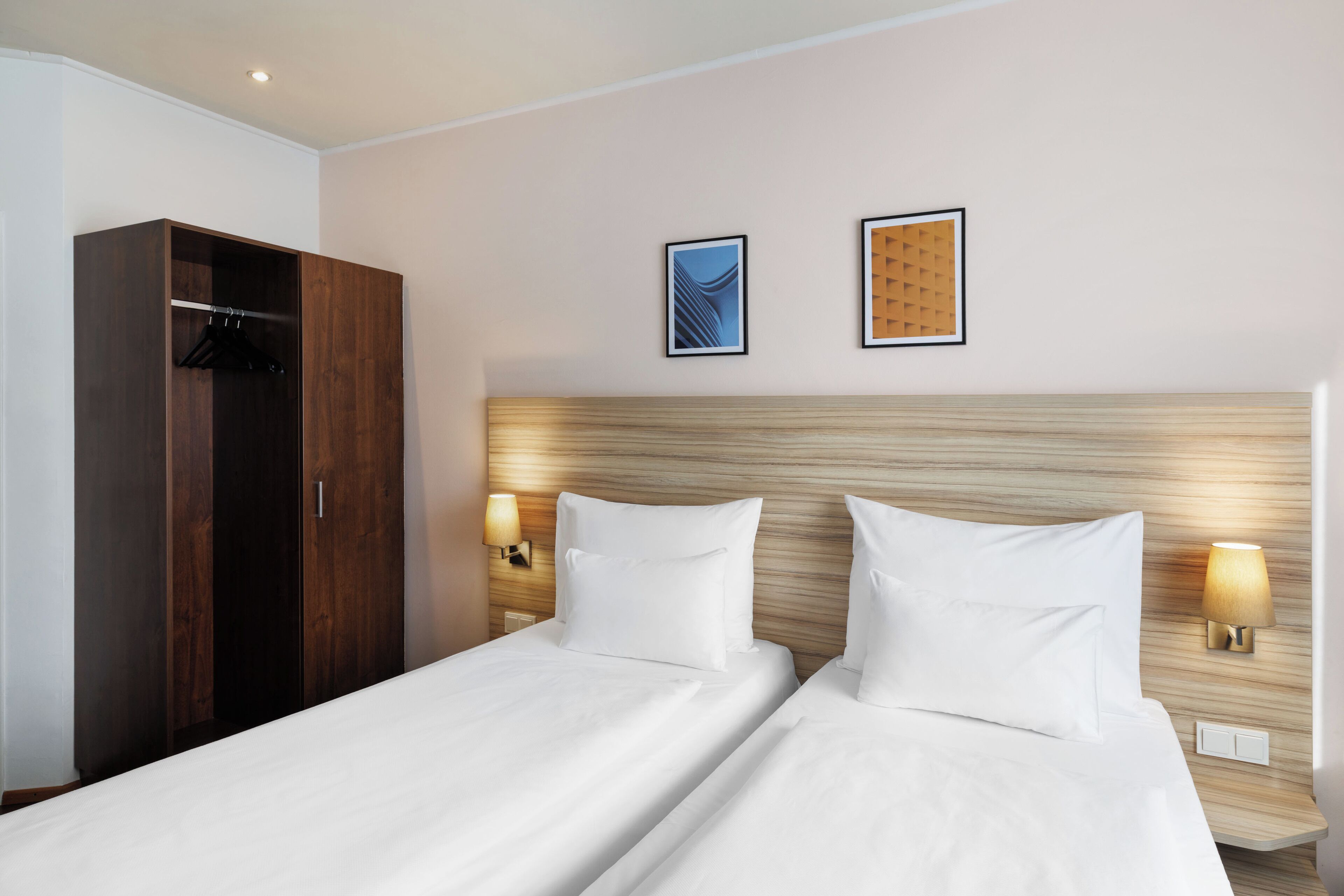A hotel room with a double bed, a wardrobe, two wall-mounted lamps, and two framed pictures on the wall.