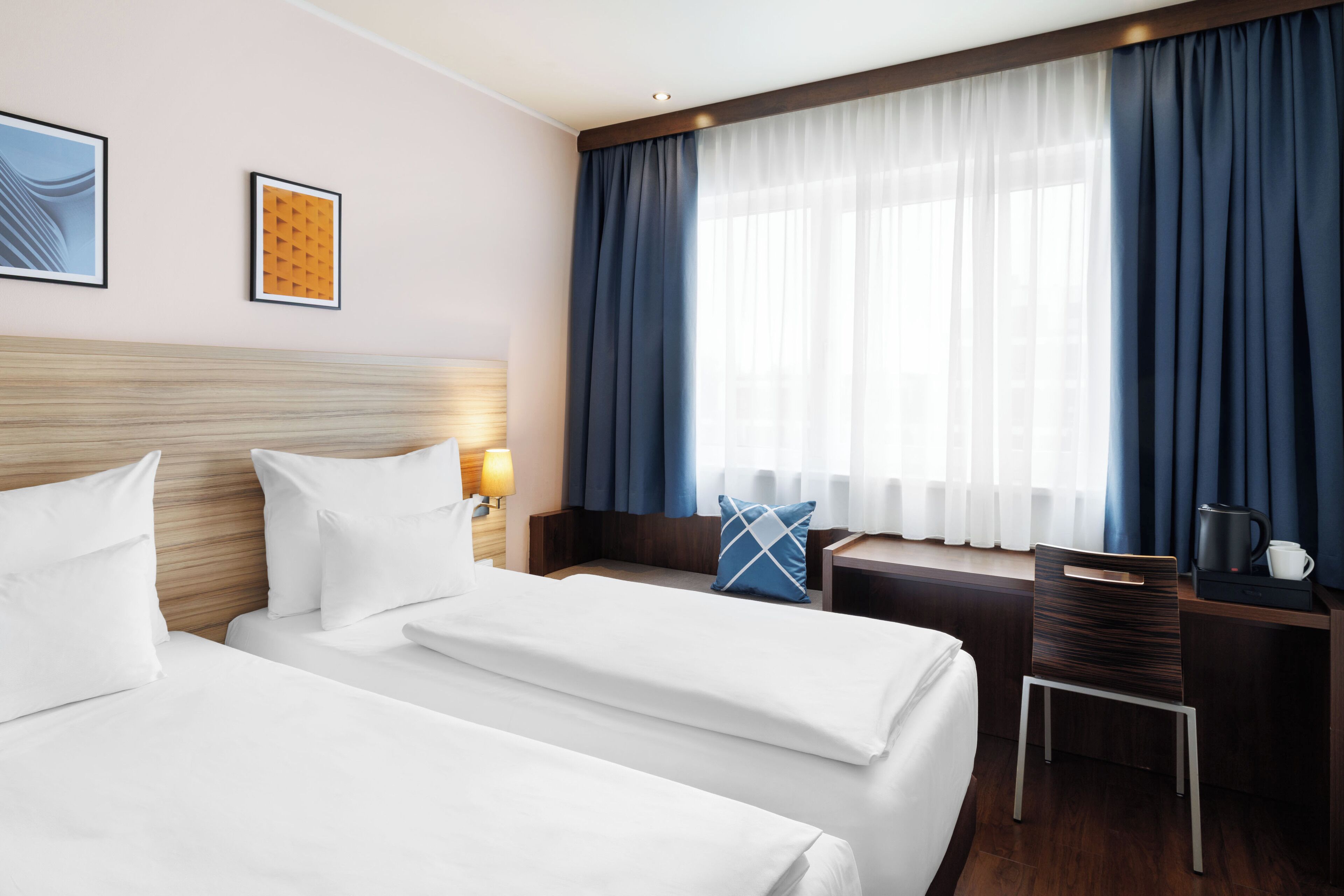 Quarto de hotel com duas camas, uma escrivaninha, uma cadeira, uma cafeteira e uma janela com cortinas.