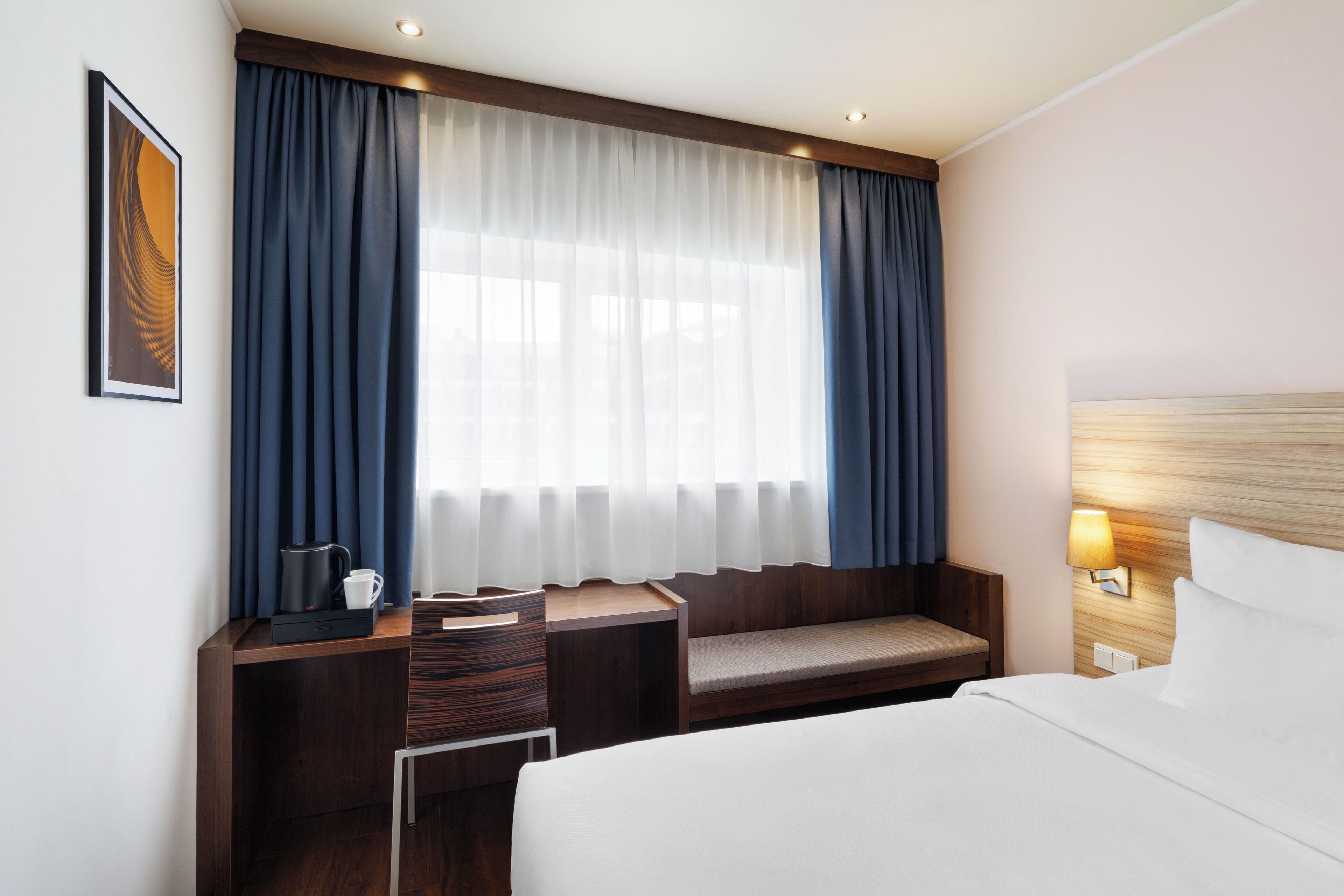 Ein Hotelzimmer mit Bett, Schreibtisch mit Kaffeemaschine, einer Bank und einem Fenster mit Vorhängen.
