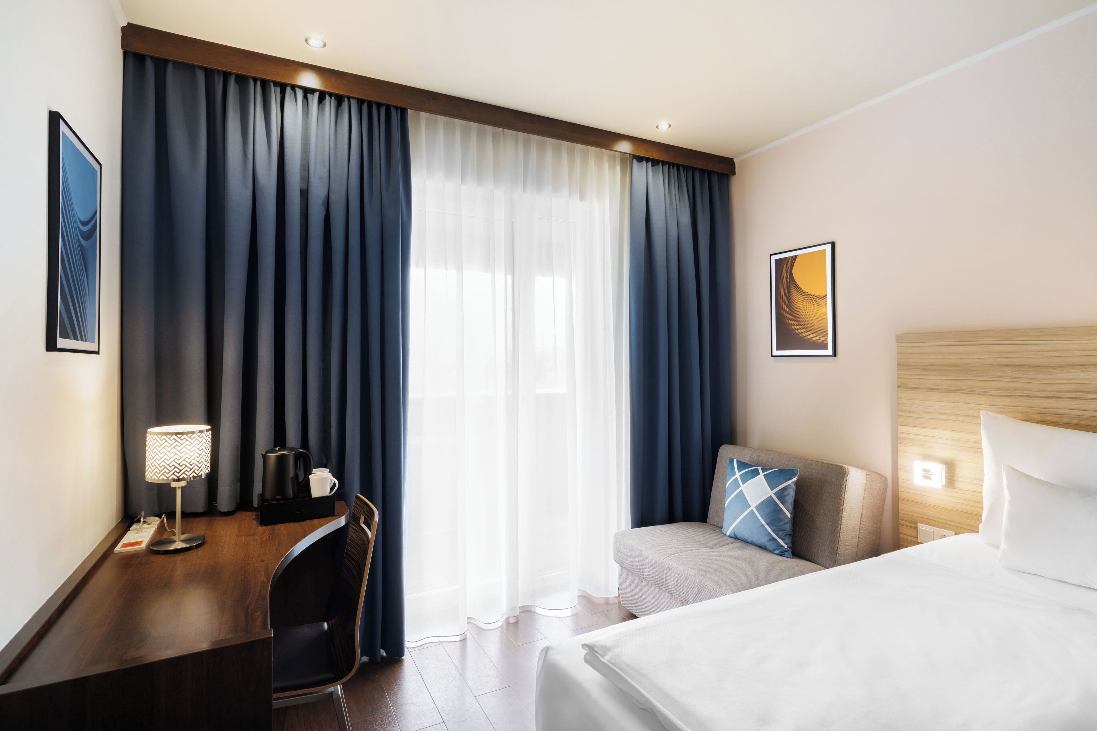 A hotel room with a bed, a desk with a lamp, a chair, and a sofa.