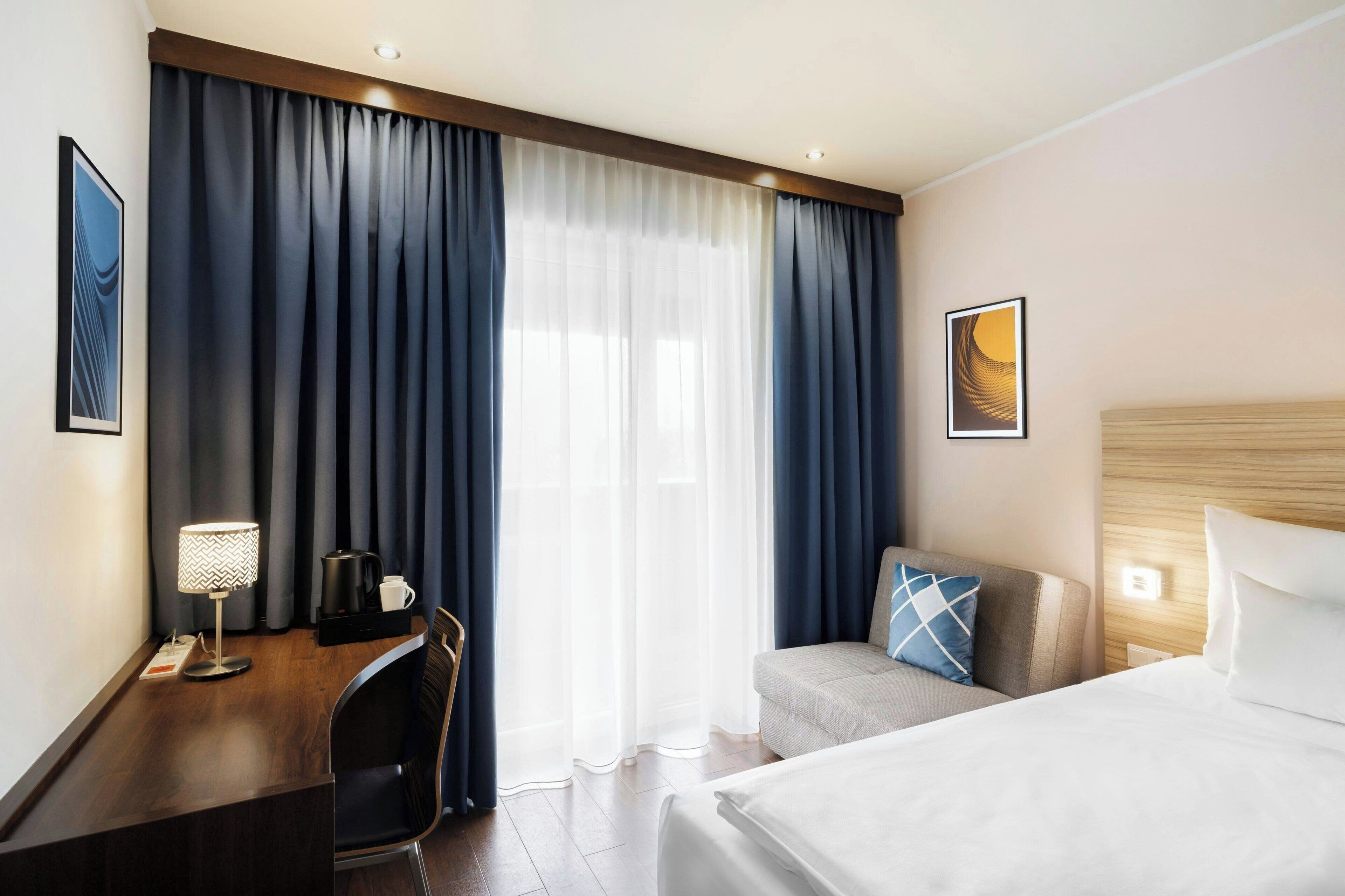 Een hotelkamer met een bed, een bureau met lamp, een stoel, een bank en een groot raam met gordijnen.