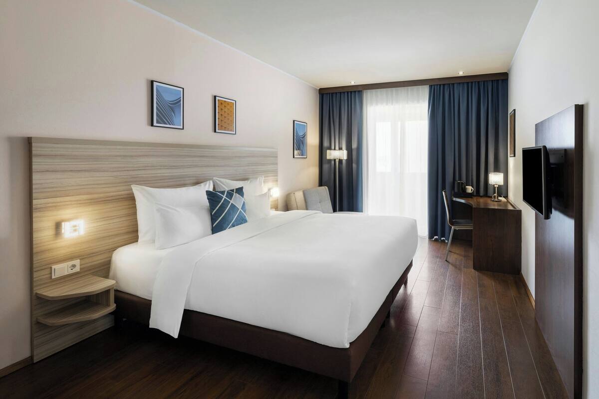 A hotel room with a large bed, bedside tables, a desk, a chair, a TV, and a window with curtains.