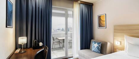 A hotel room with a bed, a desk with a lamp, a chair, a sofa, and a view of the outdoors through a sliding door.